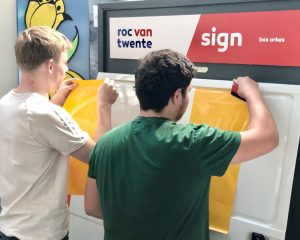 2 Studenten doen praktijkoefeningen folie plakken met rakeltechniek tijdens Appcademy workshop en gastcollege op ROC / MBO signopleiding